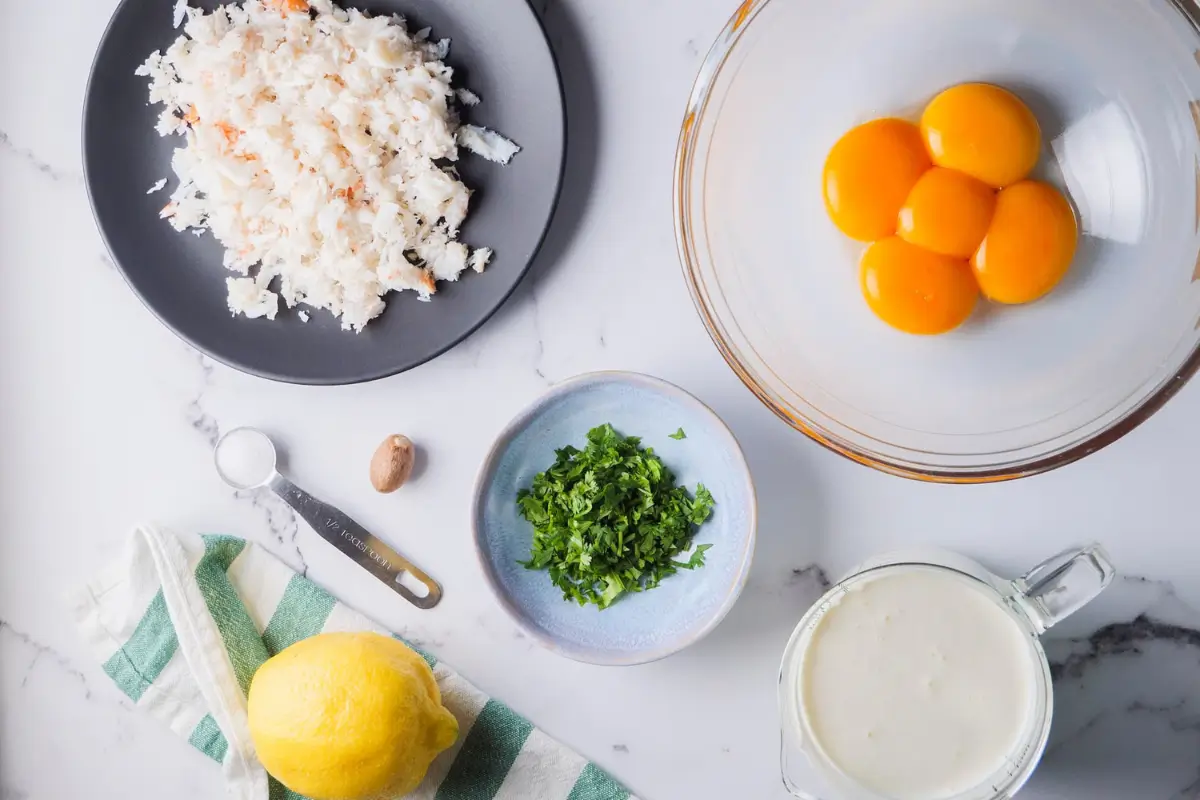 Crab Brulee Recipe Ingredients: Fresh crab meat, egg yolks, heavy cream, chives, lemon, nutmeg, and salt on a marble surface.