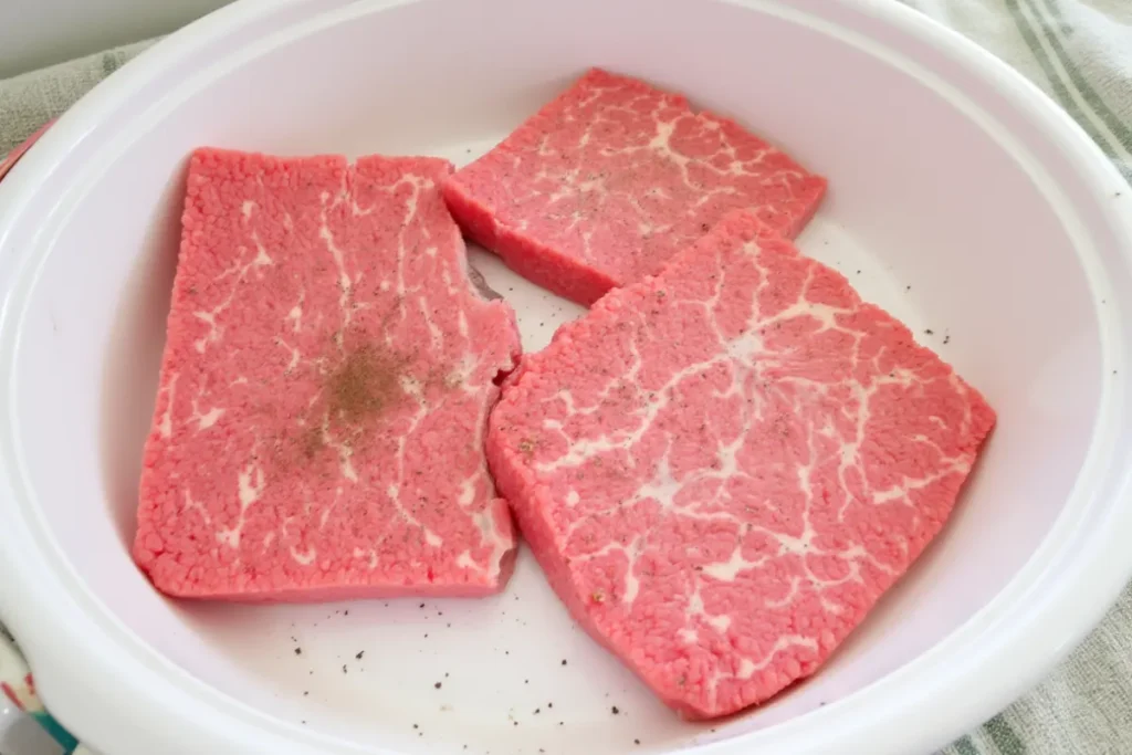 Three pieces of uncooked cube steak in a white slow cooker.