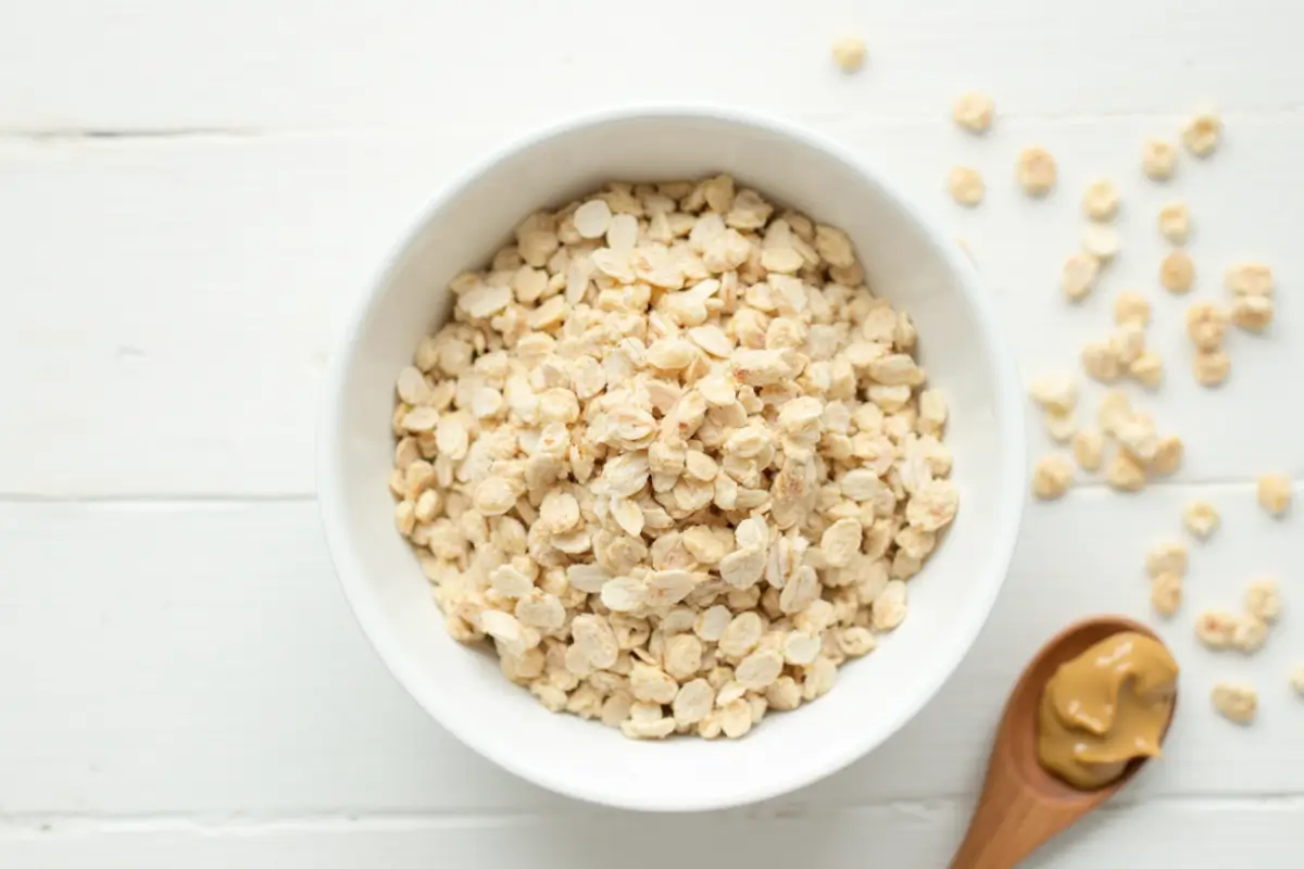 Dry rolled oats in a white bowl with a wooden spoon of peanut butter alongside, on a white wood surface.